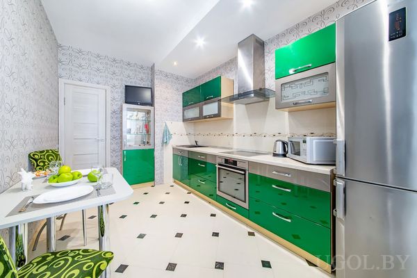 Современная кухня с зелёными и серебристыми фасадами, техникой, сервированным столом и светлым фоном.