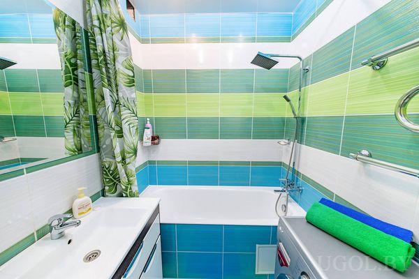Компактная ванная с плиткой в зелёно-синих тонах, ванной с занавеской, стиральной машиной и зеркалом над раковиной.