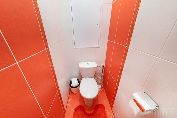 Узкий туалет с яркой плиткой, красным ковриком, мусорным ведром и белым люком над унитазом.