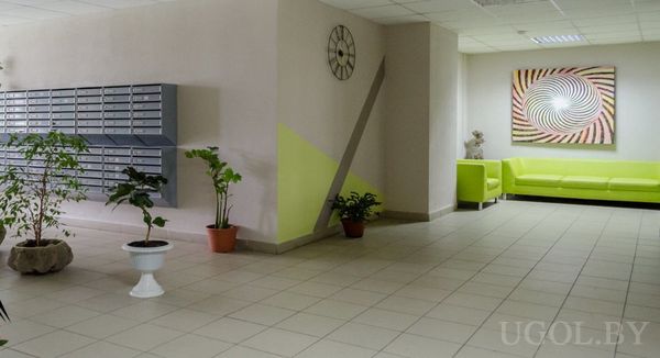 Интерьер холла с почтовыми ящиками, зелёным диваном, спиральной картиной и декоративными растениями.