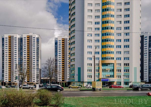 Городской пейзаж с многоэтажками с жёлто-синими вставками, улицей с машинами и деревьями на переднем плане.