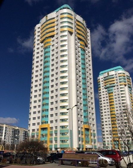 Современное жилое здание цилиндрической формы с бирюзово-жёлтыми акцентами и машинами на фоне ясного неба.