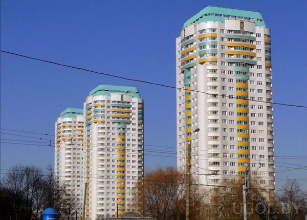 Городской вид с тремя цилиндрическими башнями, цветными фасадами и деревьями на фоне голубого неба.