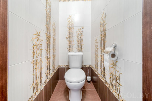 Узкое помещение с унитазом, ёршиком, держателем и плиткой с золотистым растительным орнаментом.