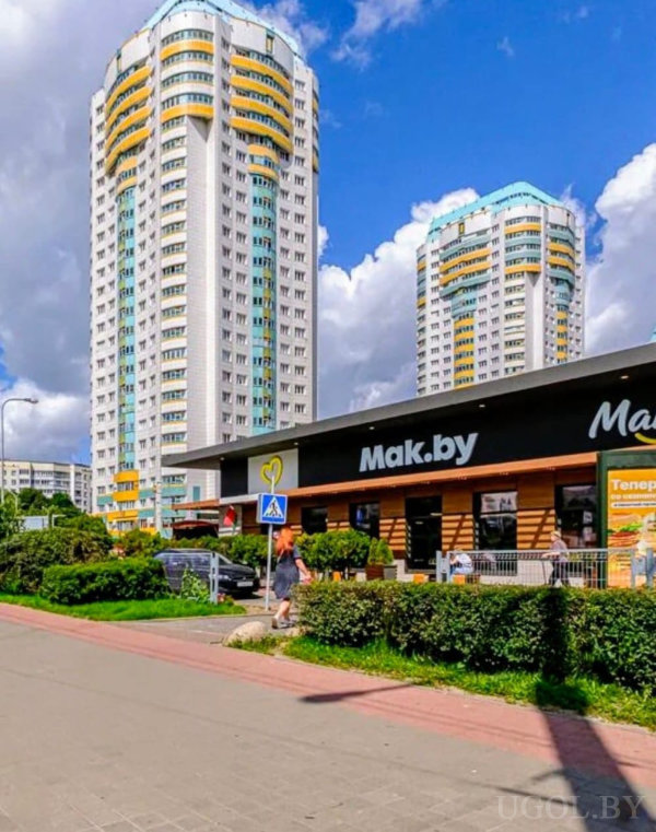 Уличная сцена с жилыми домами, магазином, пешеходной зоной, зеленью и рекламной вывеской.