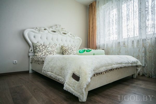 Комната с кроватью, бежевыми шторами, тюлем, деревянным полом и декоративными подушками.