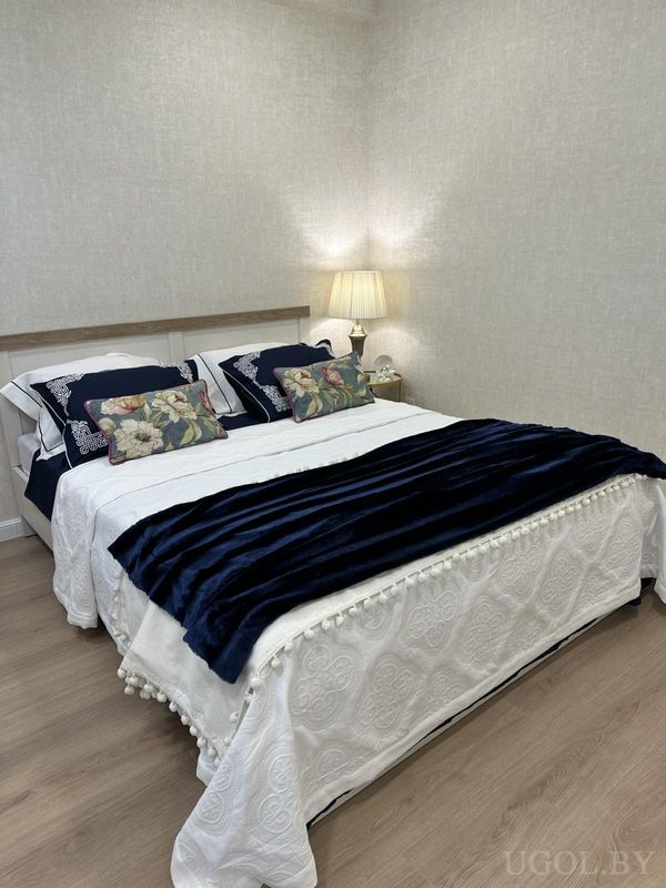 Уютная спальня с белым покрывалом, синим пледом, подушками с вышивкой и цветочным узором, рядом — тумбочка с лампой.