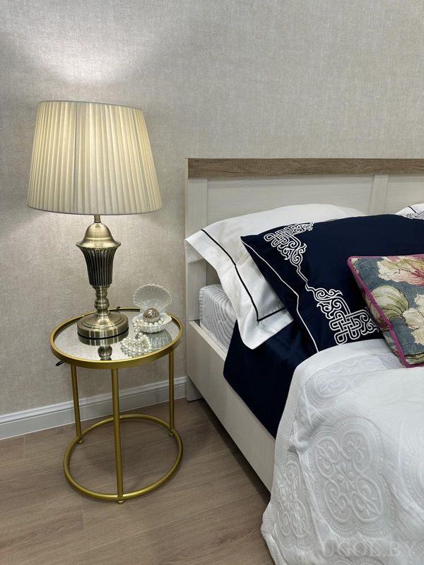 Элегантный уголок спальни с круглой тумбочкой, лампой с плиссированным абажуром, стеклянной шкатулкой и бело-синей постелью.