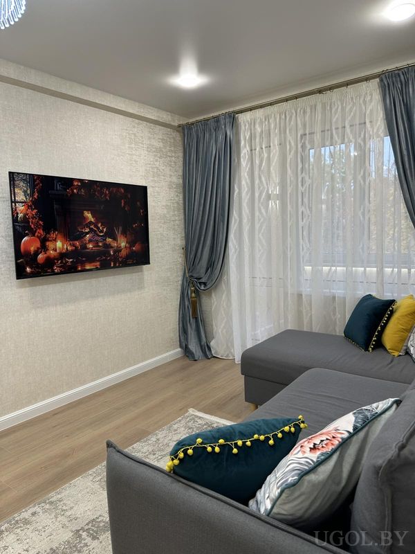 Современная гостиная с серым диваном, подушками в жёлтом, бирюзовом и цветочном стиле, телевизором с осенним камином и шторами.