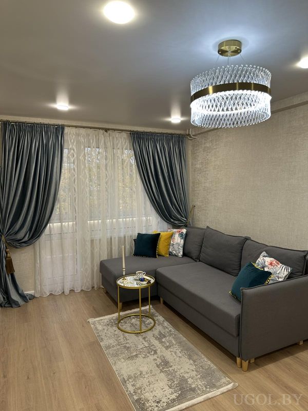 Элегантная гостиная с серым диваном, цветными подушками, круглым столиком с декором, люстрой и шторами в тёмно-бирюзовом цвете.
