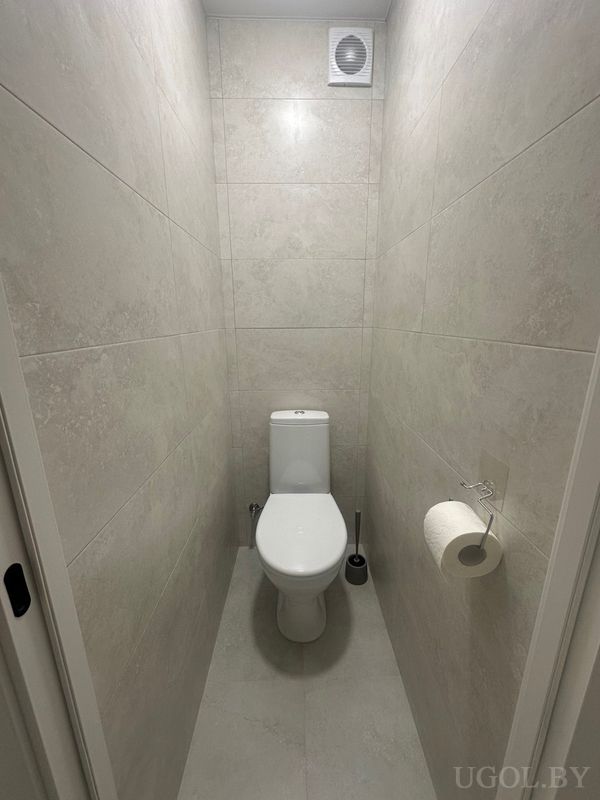 Узкий туалет с белым унитазом, туалетной бумагой на стене, вентиляцией и плиткой с мраморным рисунком.