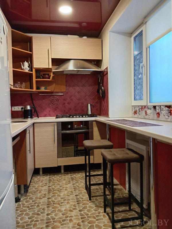 U-образная кухня с деревянными фасадами, красный фартук, барная зона с табуретами и глянцевый потолок.