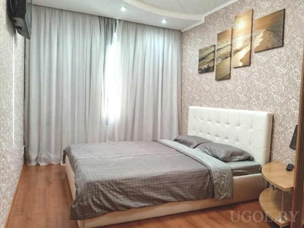 Большая кровать с белым изголовьем и серым бельём, стена с обоями и пятичастным панно с морским пейзажем, деревянный пол, прикроватная тумбочка и настенное ТВ напротив.