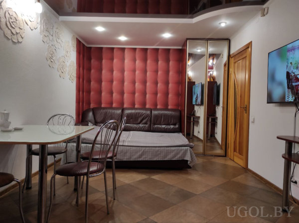  Коричневый кожаный диван-кровать с серым покрывалом на фоне красной мягкой панели, стеклянный стол с металлическими стульями, настенное ТВ, зеркальный шкаф и потолок с точечным светом.