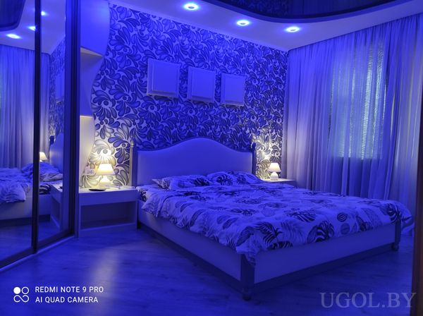 Кровать с цветочным покрывалом, синяя LED-подсветка, зеркальный шкаф, тумбочки с лампами и декоративная стена с панно.