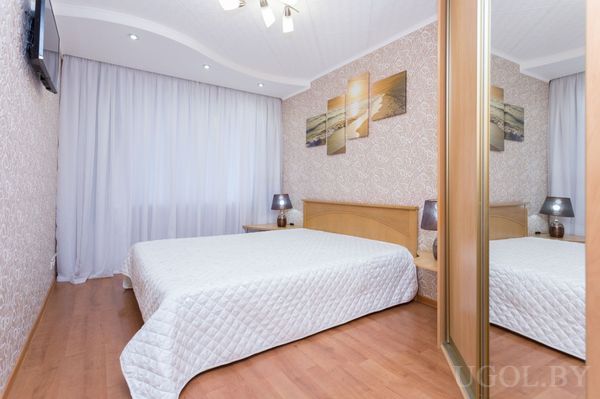 Кровать с белым покрывалом, тумбы с лампами, панорамное панно над изголовьем, телевизор и зеркальный шкаф.