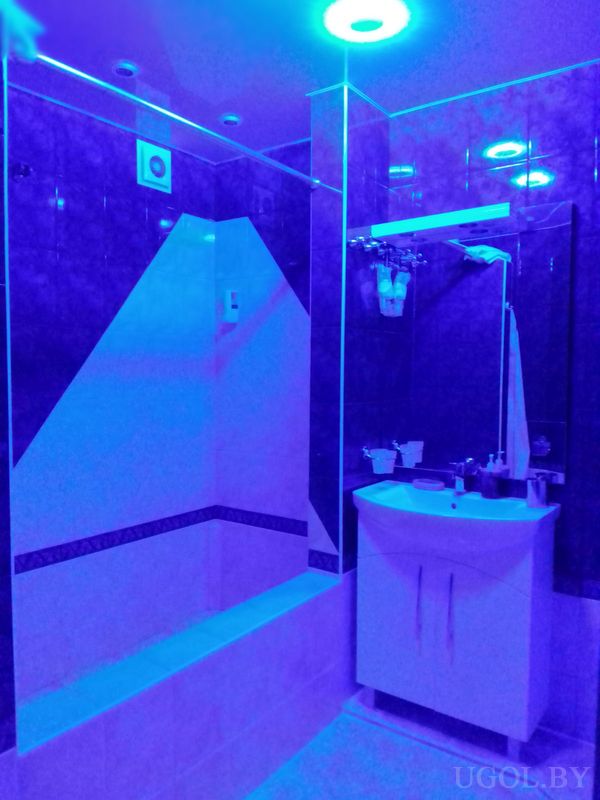 Тёмная плитка, душ с геометричным узором, белая тумба с раковиной, зеркало и синяя подсветка на потолке.