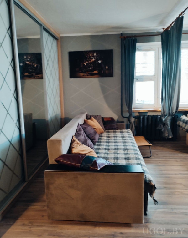 Уютная гостиная с бежевым диваном, цветными подушками, клетчатым пледом и зеркальными дверями шкафа, создающими эффект глубины.