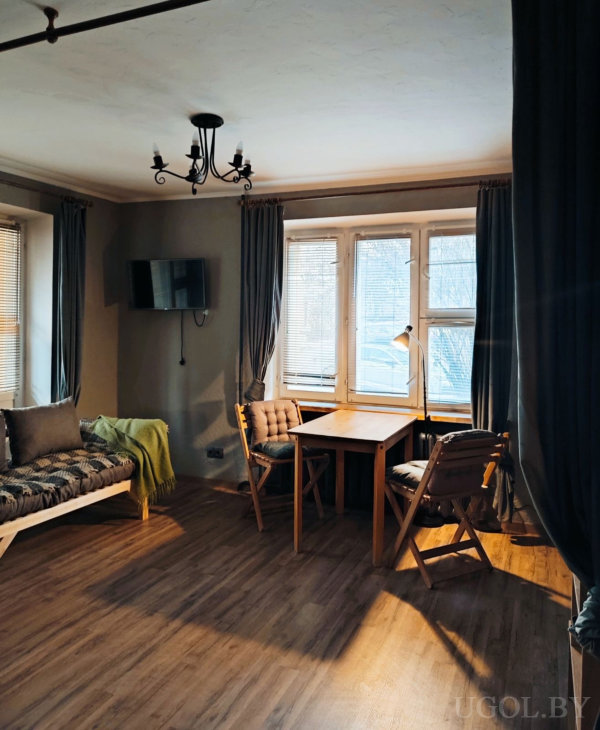 Светлая комната с деревянным столом и креслами у окна, мягкой скамьей, телевизором и люстрой в стиле ретро.