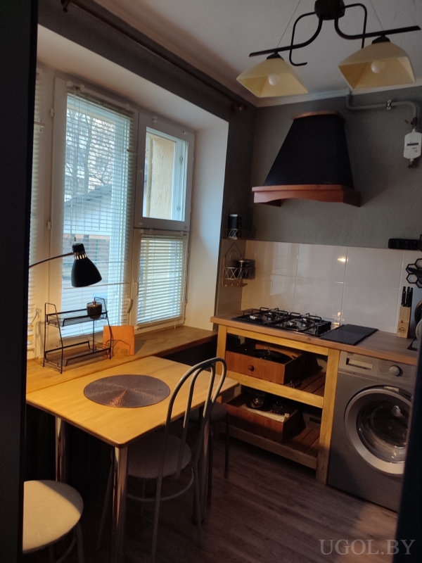 Компактная кухня с плитой, вытяжкой, стиральной машиной и обеденной зоной у окна с настольной лампой.