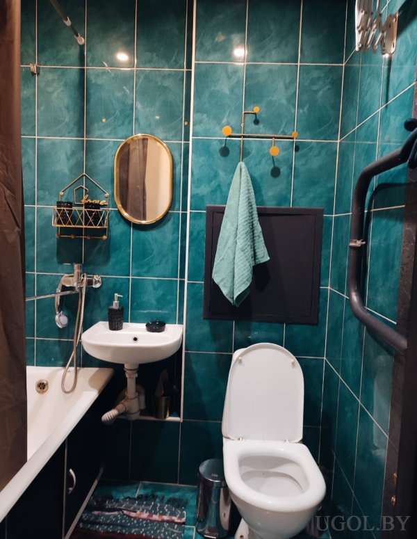 Небольшая ванная с белой сантехникой, золотым зеркалом, декоративной вешалкой и чёрным полотенцесушителем.