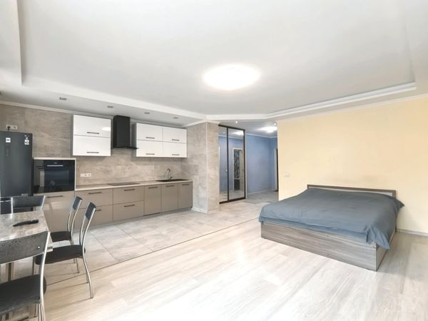 Открытая студия с кухней вдоль стены, столом на четыре персоны, серой кроватью и зеркальным шкафом в коридоре.