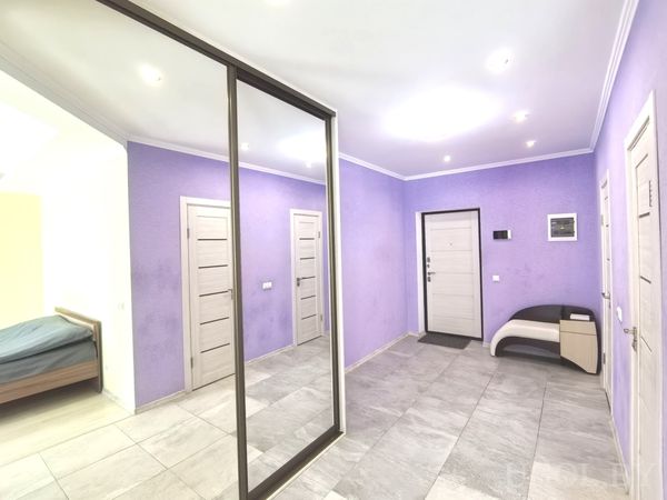 Светлый коридор с сиреневыми стенами, белыми дверями, зеркальным шкафом и скамьёй у входа.