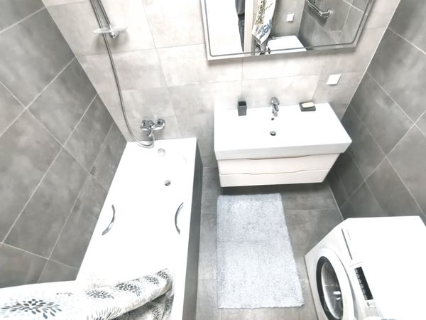 Функциональная ванная с серой плиткой, белой сантехникой, стиральной машиной и занавеской с листовым узором.