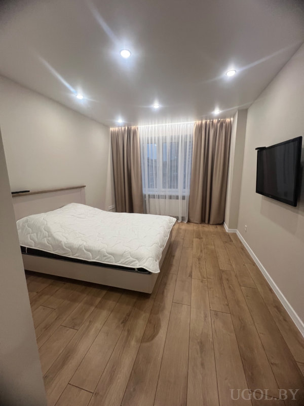 Минималистичная спальня с белым матрасом, настенным телевизором и большими окнами с бежевыми шторами, оформлена в спокойных тонах.