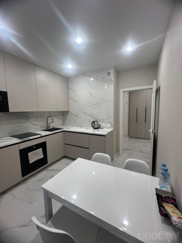 Минималистичная кухня с белым столом, закусками, встроенной техникой и открытой дверью в коридор, оформлена в теплых оттенках.