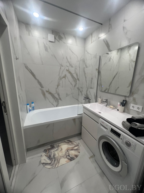 Современная ванная с белой ванной, стеклянной перегородкой, встроенной стиральной машиной и мраморной плиткой.