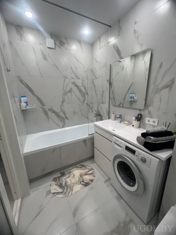 Светлая ванная с зеркалом, стиральной машиной, стеклянной перегородкой и декоративным ковриком, выполнена в мраморной гамме.