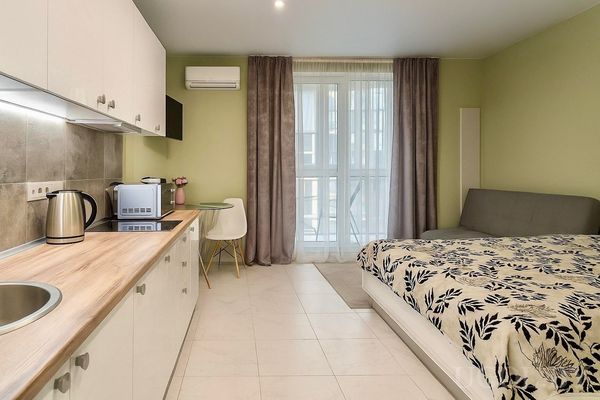 Комната с кухонной зоной, столом с вазой, кроватью с чёрно-белым текстилем и окнами с серыми шторами.