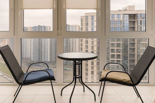 Застеклённая зона с двумя креслами, круглым столом, белой плиткой и видом на высотки через большие окна.