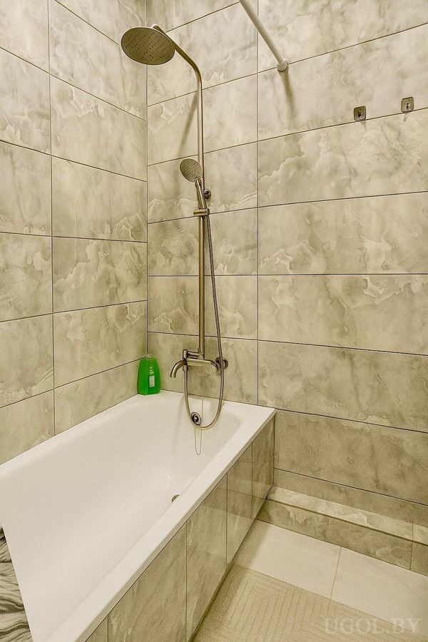 Интерьер с ванной, душевой стойкой, серой плиткой с мраморным узором и ковриком у края ванны.