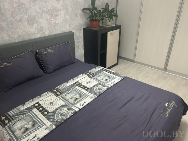 Уютная кровать с фиолетовым покрывалом и подушками с надписью «Good Sleep», декоративный плед с сердечками и надписями, рядом тумба с растениями.