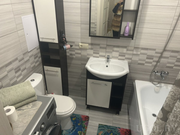 Белая ванна, раковина, шкафы и зеркало, стиральная машина с полотенцами, плитка серых оттенков и яркие коврики.