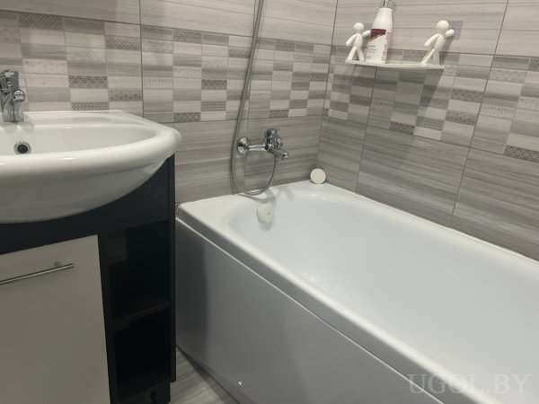 Белая ванна и раковина, тёмный шкаф, серо-белая плитка и полка с фигурками и шампунем.