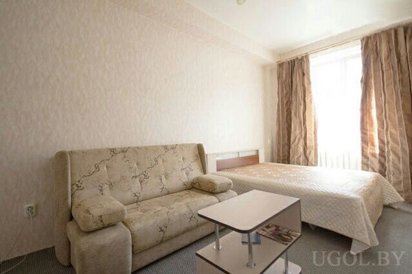 Небольшая спальня с бежевым диваном с цветочным узором, двуспальной кроватью со светлым покрывалом и журнальным столиком.