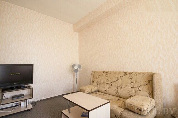 Комната с бежевым диваном, журнальным столиком, телевизором на тумбе и напольным вентилятором.