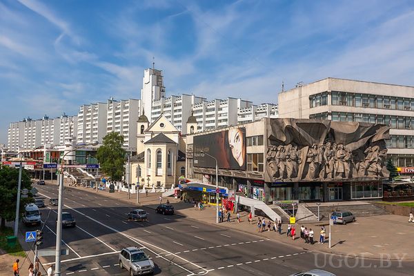 Улица с белой церковью, современными зданиями, большим барельефом на фасаде и оживлённым движением.