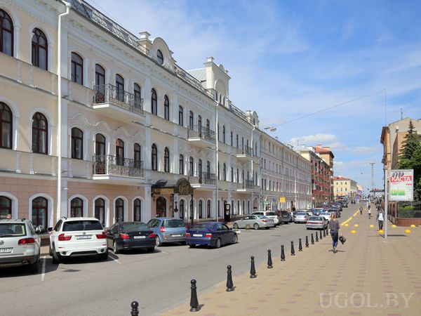Городская улица с историческими домами с арочными окнами и балконами, припаркованными машинами и пешеходами.
