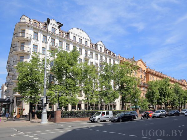 Угловое здание с белыми фасадами, балконами с чёрными перилами, деревьями вдоль тротуара и оживлённой улицей.