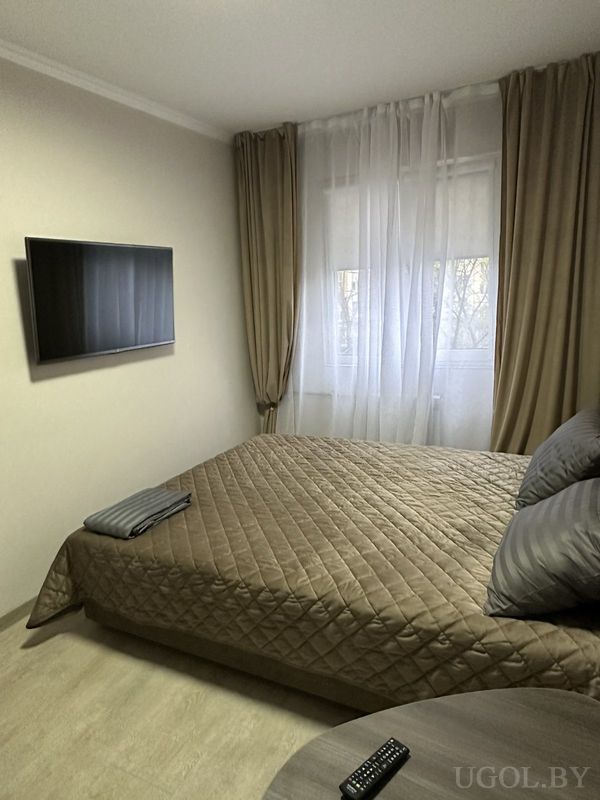 Современная спальня с коричневым текстилем, телевизором на стене, тюлем и шторами, на прикроватной тумбе — пульт.