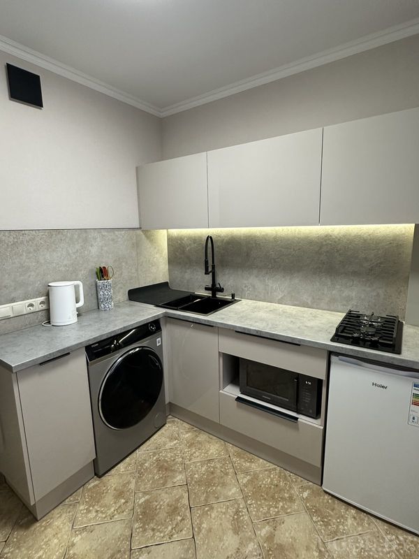 Компактная кухня с серым фартуком, чёрной мойкой, техникой под столешницей и светлыми фасадами без ручек.