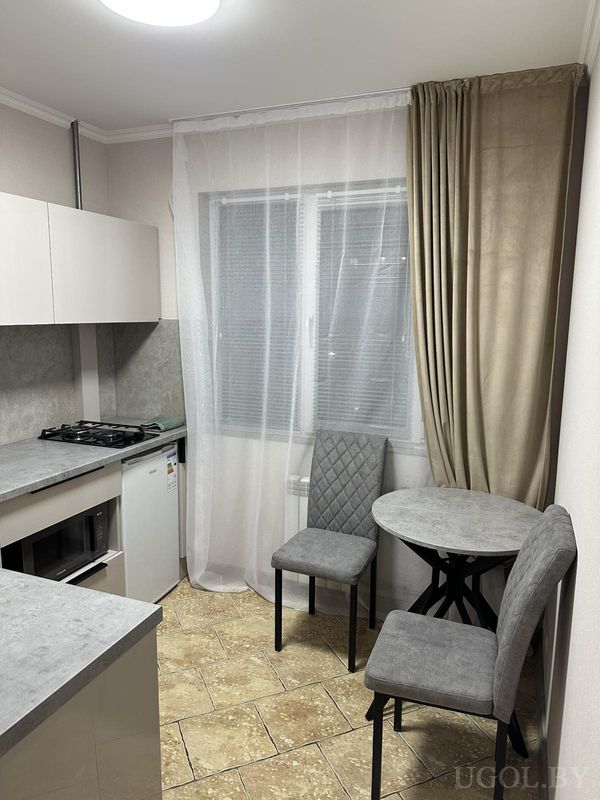 Небольшая кухня с круглым столом, двумя стульями с ромбовидной строчкой, плитой и окном с тюлем и жалюзи.