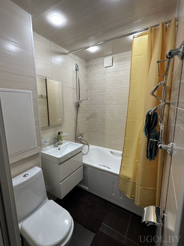 Функциональная ванная с белой сантехникой, жёлтой занавеской, зеркалом над раковиной и тёмным ковриком на полу.