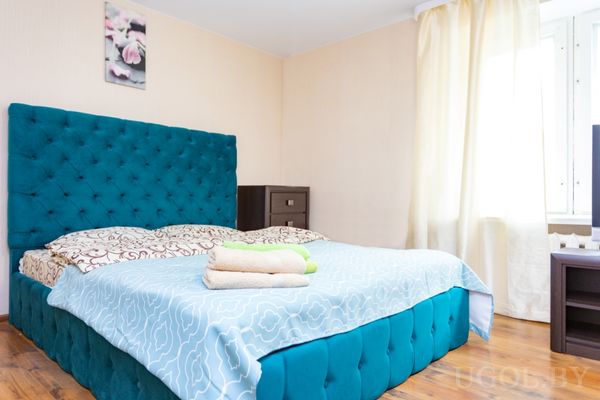 Современная спальня с кроватью в бирюзовом изголовье, светлым покрывалом, подушками и сложенными полотенцами.