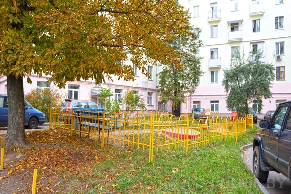 Небольшая детская площадка с песочницей и лавочкой, окружённая жилыми домами и деревьями.