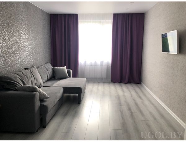 Комната с угловым диваном, телевизором на стене, светлым ламинатом и окнами с тюлем и плотными шторами.